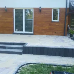 Nowoczesny dom z elewacją imitującą drewno, taras wyłożony jasną kostką brukową, schody z ciemnych elementów, widoczne lampy zewnętrzne i okno.