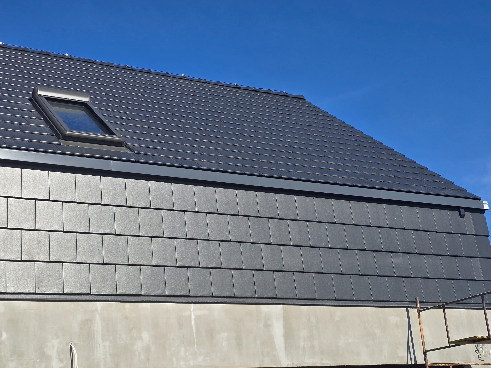 Fragment dachu z czarnej dachówki bitumicznej w kształcie prostokątnych płytek, z oknem dachowym, na tle błękitnego nieba, widoczne rusztowanie.