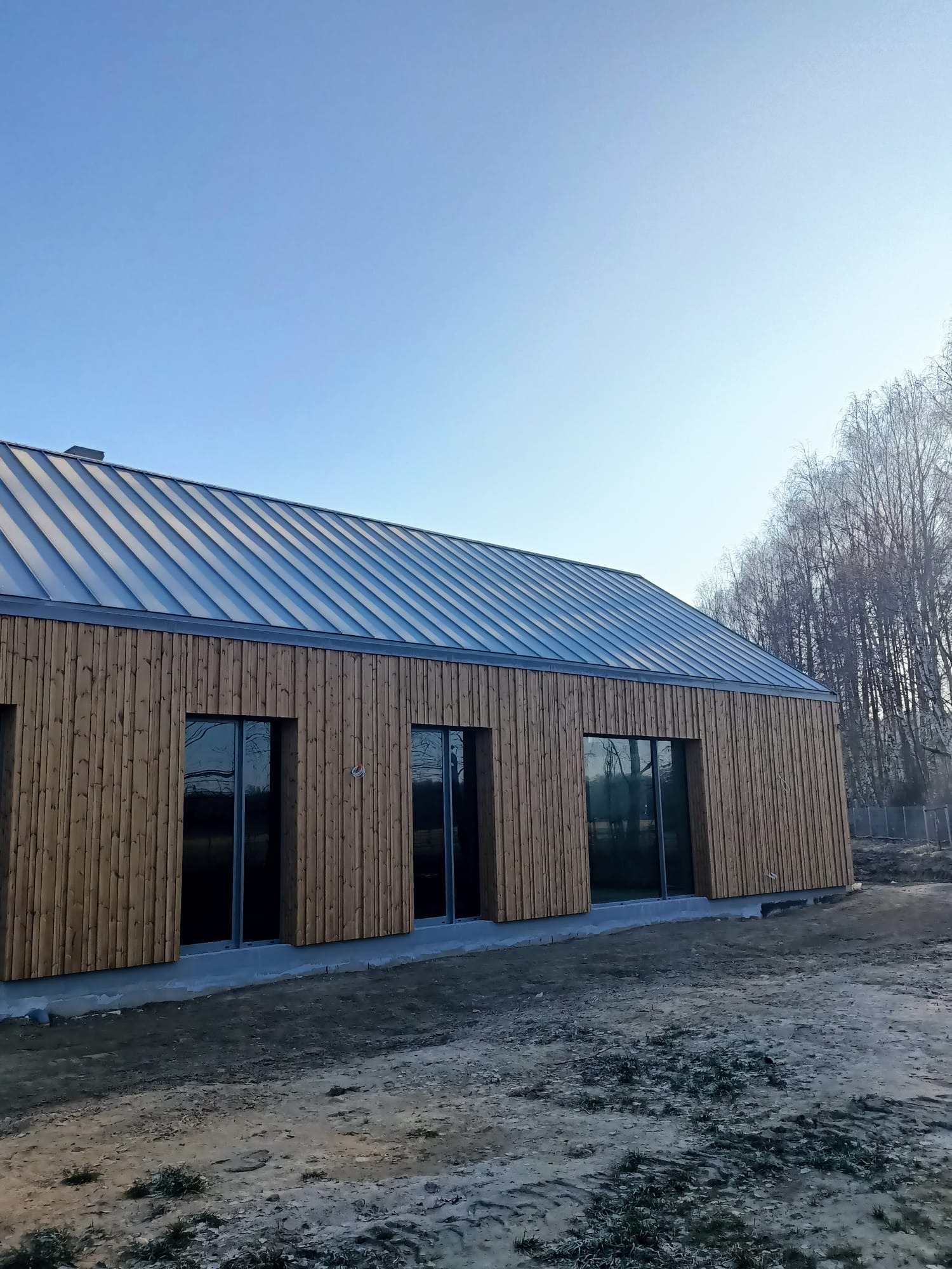 Nowoczesny dom jednorodzinny z drewnianą elewacją i metalowym dachem na etapie wykończenia, widoczne okna z ciemnymi ramami i surowy teren wokół budynku.