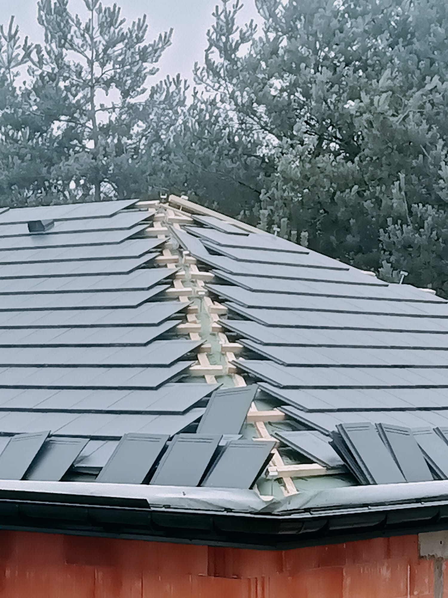 Montaż grafitowej dachówki ceramicznej na drewnianej konstrukcji dachu, widok z dołu na tle szarych drzew iglastych.