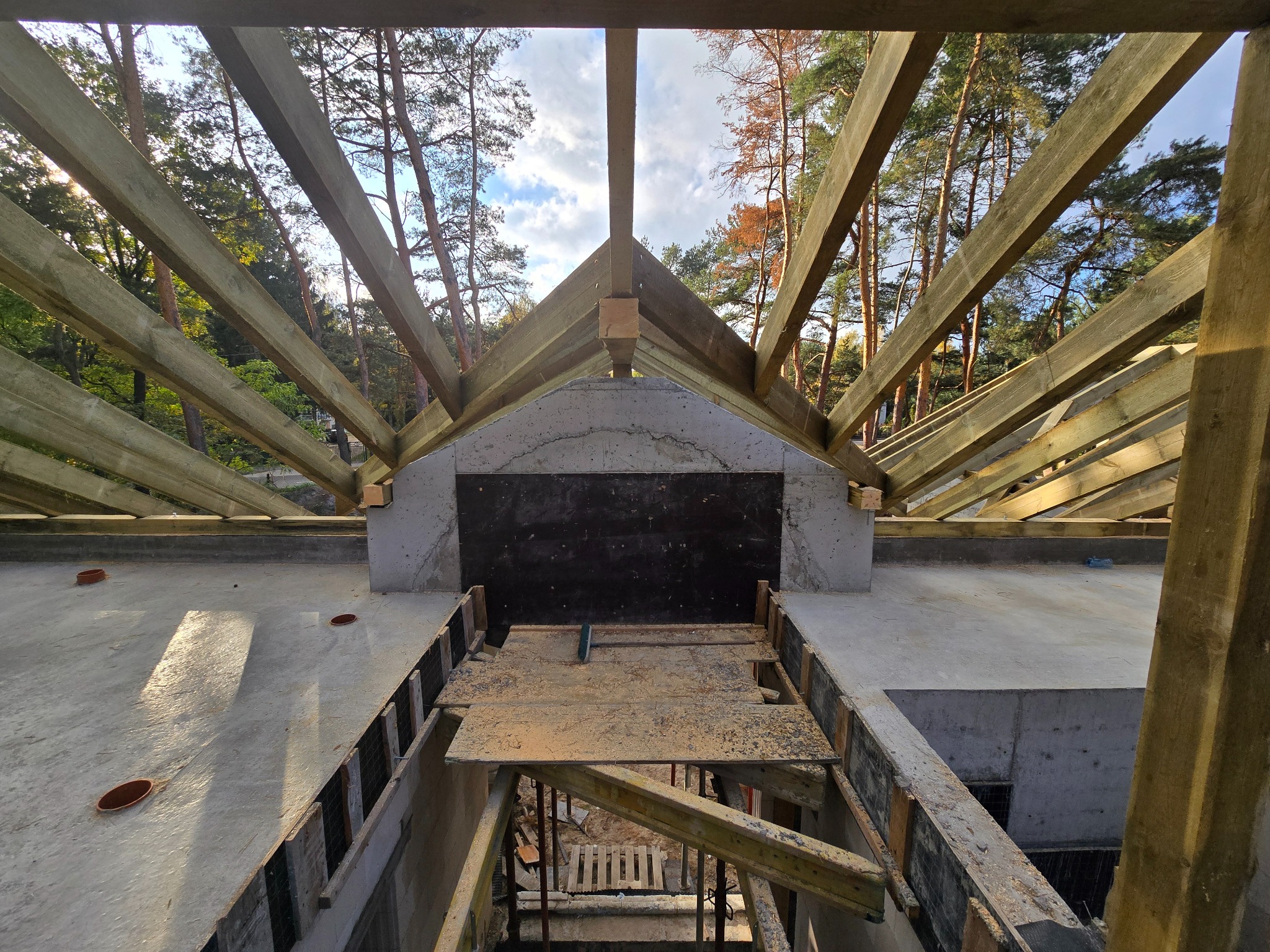 Surowa konstrukcja domu w trakcie budowy: widok na drewniany szkielet dachu z betonową ścianą szczytową na tle lasu i błękitnego nieba.