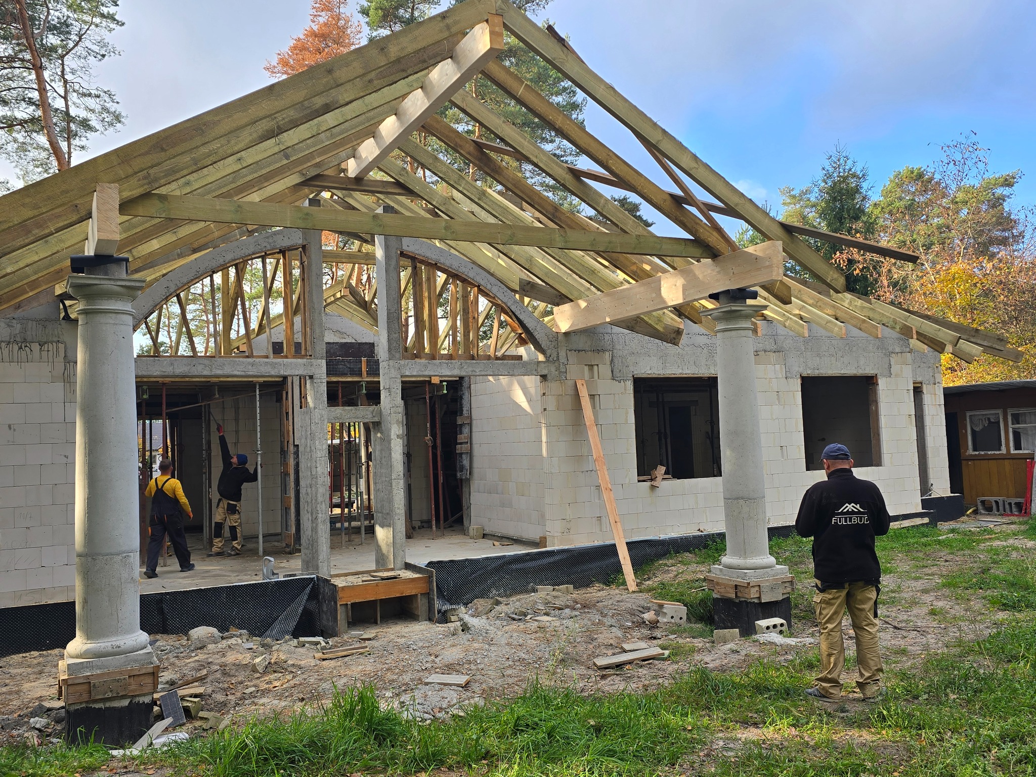 Budowa domu z białych bloczków, widoczne drewniane elementy konstrukcji dachu, dwie kolumny z dekoracyjnymi głowicami, dwóch pracowników w środku budynku, trzeci obserwuje z zewnątrz, na ubraniu...