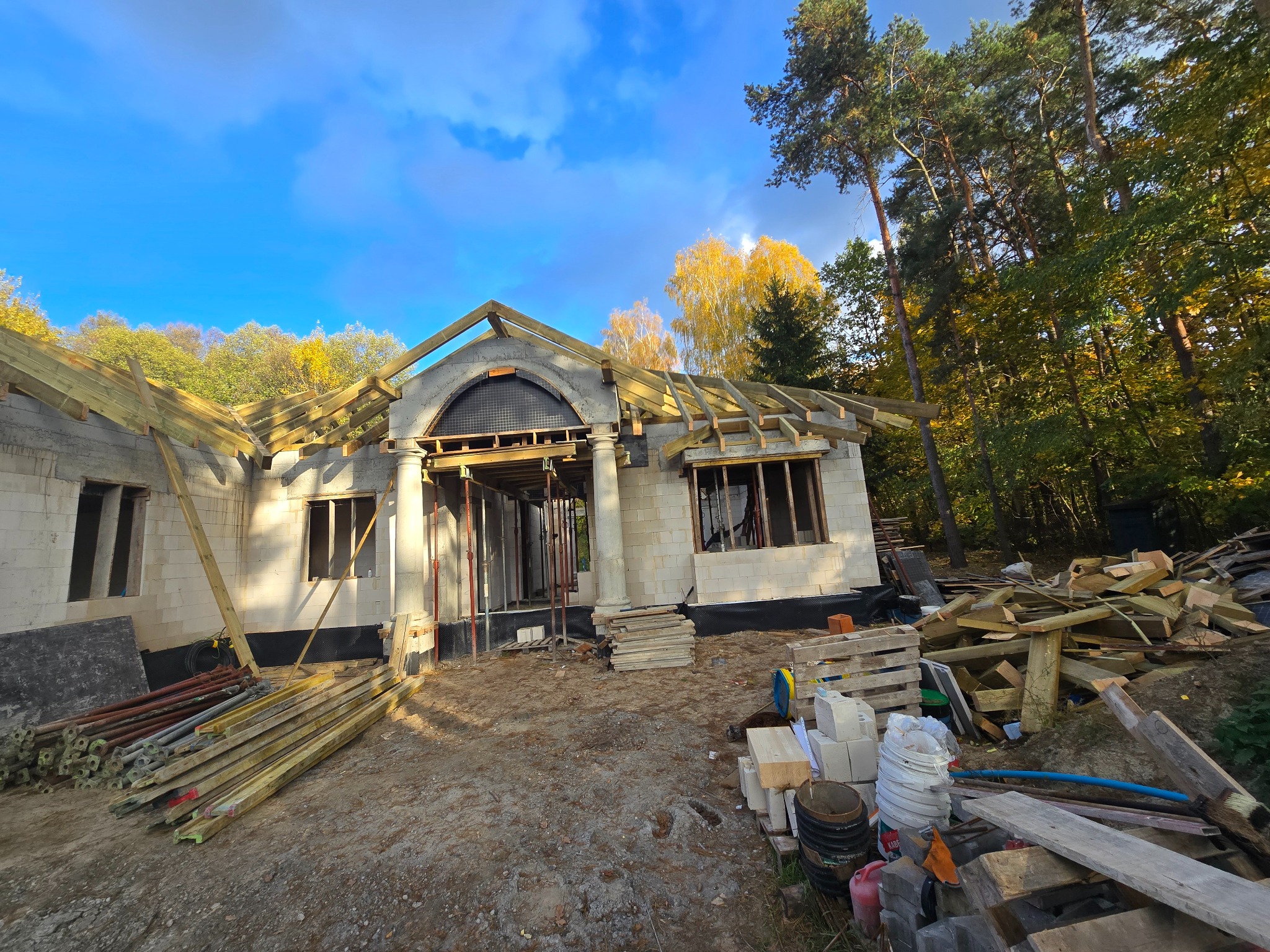 Budowa domu jednorodzinnego z elementami łukowymi nad wejściem, widoczne drewniane elementy konstrukcji dachu i ściany z białych bloczków, w tle las i błękitne niebo z chmurami.