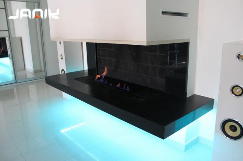 Nowoczesny biokominek z czarnym obramowaniem, podświetlony niebieskim światłem LED, płomienie widoczne w palenisku, logo firmy widoczne w rogu, minimalistyczne wnętrze.