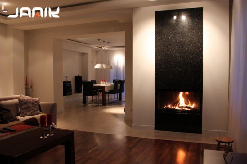 Nowoczesny kominek z czarnym, błyszczącym obramowaniem z kamienia, płomienie widoczne w palenisku, otwarte wnętrze domu w tle z jadalnią i salonem.