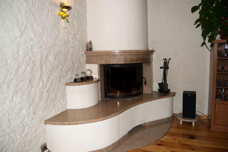 Nowoczesny narożny kominek z obudową z beżowego kamienia i białymi elementami, z akcesoriami kominkowymi po prawej stronie i dekoracyjnymi kulami po lewej, na tle teksturowanej ściany.