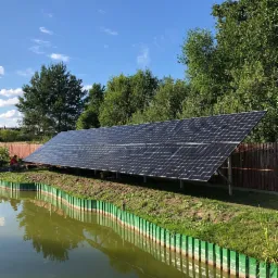 Długi panel słoneczny zamontowany nad brzegiem stawu, odbicie zieleni w wodzie, pracownik w czerwonej koszulce siedzi obok, błękitne niebo z białymi chmurami w tle.