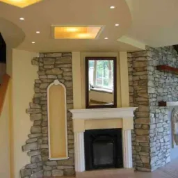 Aranżacja wnętrza z dekoracyjnym kamieniem na ścianach, kominkiem z białym obramowaniem i czarnym wkładem, oraz podwieszanym sufitem z oświetleniem punktowym i podświetlanymi panelami.