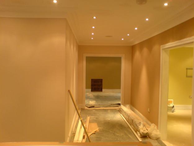 Korytarz w trakcie prac wykończeniowych, widoczne świeżo położone ściany w odcieniach beżu, surowa posadzka, listwy przypodłogowe w folii, oświetlenie halogenowe w suficie i otwarte przejście...