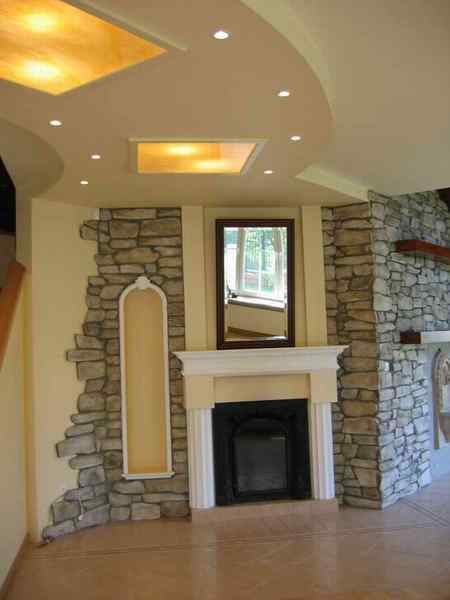 Aranżacja wnętrza z dekoracyjnym kamieniem na ścianach, kominkiem z białym obramowaniem i czarnym wkładem, oraz podwieszanym sufitem z oświetleniem punktowym i podświetlanymi panelami.