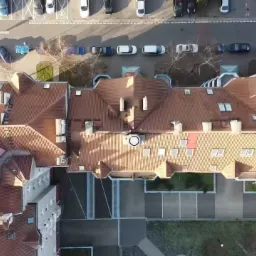 Widok z drona na rozległy budynek z czerwonym dachem, otoczony parkingiem i zielenią, w słoneczny dzień.