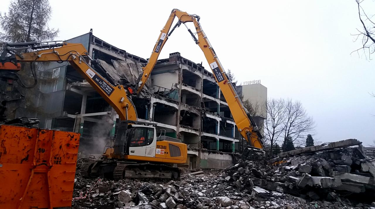 Dwie żółte koparki Liebhherr 936 z długimi ramionami rozbierają wielopiętrowy budynek; gruzowisko w pierwszym planie; pochmurna pogoda.