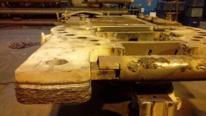 Metalowa konstrukcja z widocznymi śladami spawania i zużycia, prawdopodobnie element większej instalacji przemysłowej, z licznymi otworami i nierównościami powierzchni, oświetlona sztucznym światłem.