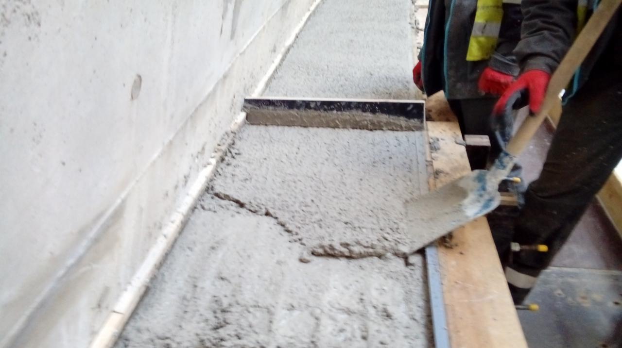 Wylewka betonowa wyrównywana szpachlą i łopatą przez pracownika w czerwonych rękawicach, widoczne szalunki i fragment ściany.