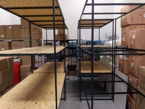 Magazyn z metalowymi regałami z płytami OSB, częściowo wypełniony kartonami i towarem tekstylnym w tle.