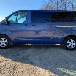 Niebieski van Ford Transit z przyciemnianymi szybami, zaparkowany na piaszczystym podłożu z widocznymi śladami opon, na tle błękitnego nieba i drzew bez liści.