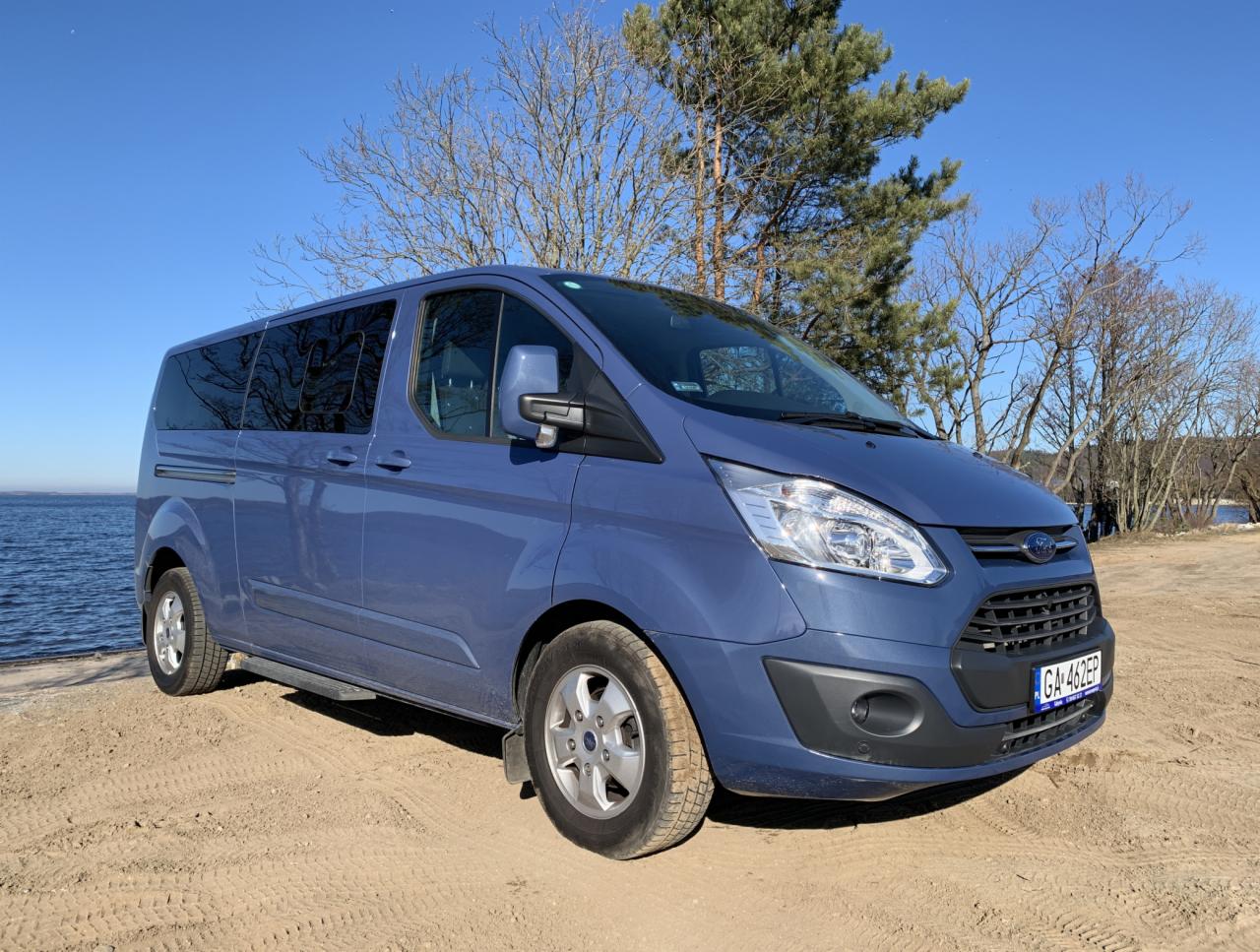 Niebieski Ford Transit Custom z przyciemnianymi szybami, zaparkowany na piaszczystym terenie w pobliżu zbiornika wodnego, z widocznym drzewem w tle i błękitnym niebem.