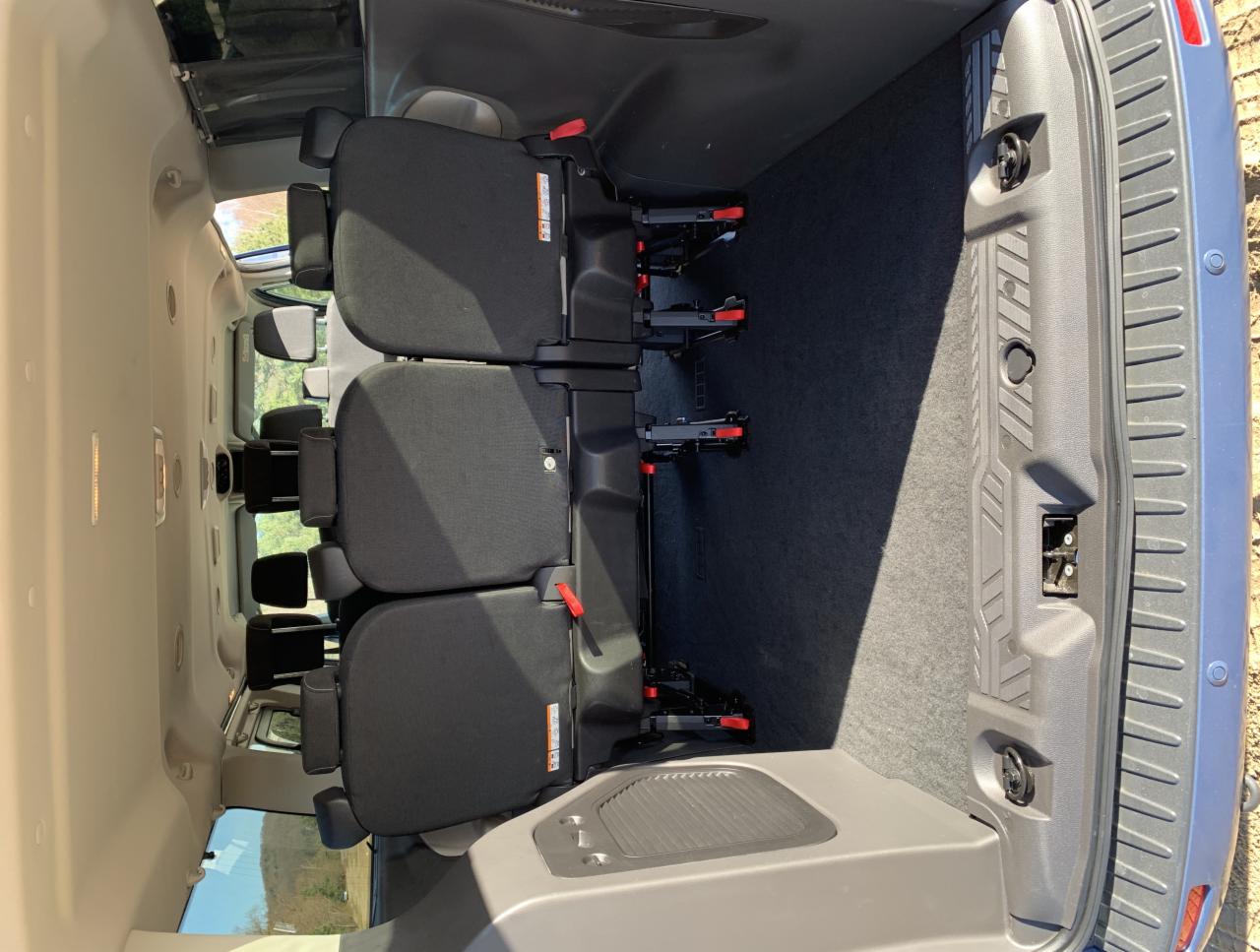 Wnętrze busa z trzema rzędami siedzeń z czarną tapicerką, widok od tyłu pojazdu, z widoczną przestrzenią bagażową wyłożoną szarym dywanem.