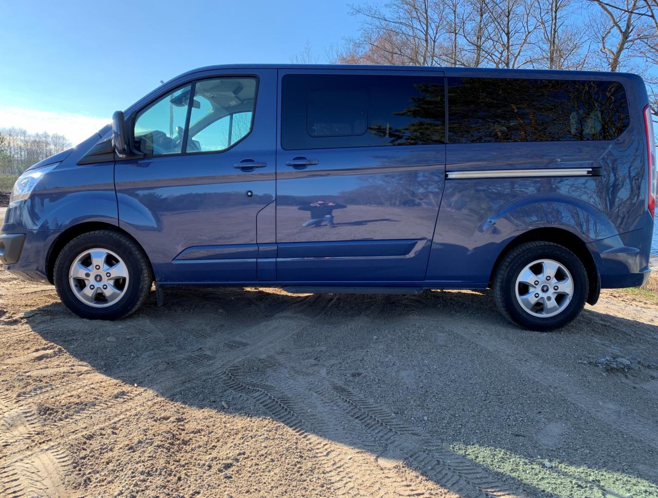 Niebieski van Ford Transit z przyciemnianymi szybami, zaparkowany na piaszczystym podłożu z widocznymi śladami opon, na tle błękitnego nieba i drzew bez liści.