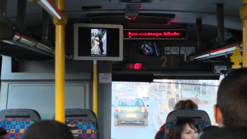 Wnętrze autobusu miejskiego z widocznym ekranem LCD wyświetlającym parę młodą, zegarem wskazującym godzinę 5:02 i komunikatem 'bez ważnego biletu' nad przednią szybą.