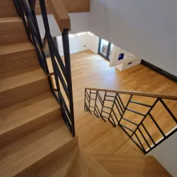 Kątowe ujęcie drewnianych schodów z czarną, geometryczną balustradą, widok z góry na jasne, drewniane podłogi w nowoczesnym wnętrzu.