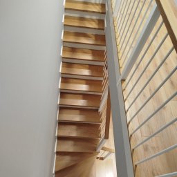 Usługi Stolarskie - Jasne, drewniane stopnie schodów z białymi podstopnicami, widziane z góry, z metalową balustradą po prawej stronie. Minimalistyczny design, naturalne światło.
