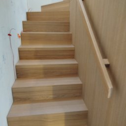 Usługi Stolarskie - Jasne, drewniane stopnie schodów z prostą balustradą, widoczne przewody na ścianie, sugerujące etap wykończeniowy. Minimalistyczny design.