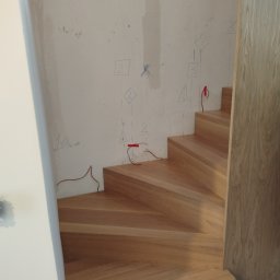 Usługi Stolarskie - Jasne, drewniane schody spiralne w trakcie montażu, widoczne oznaczenia na ścianie i wystające przewody elektryczne. Ujęcie z dołu, podkreślające kształt.