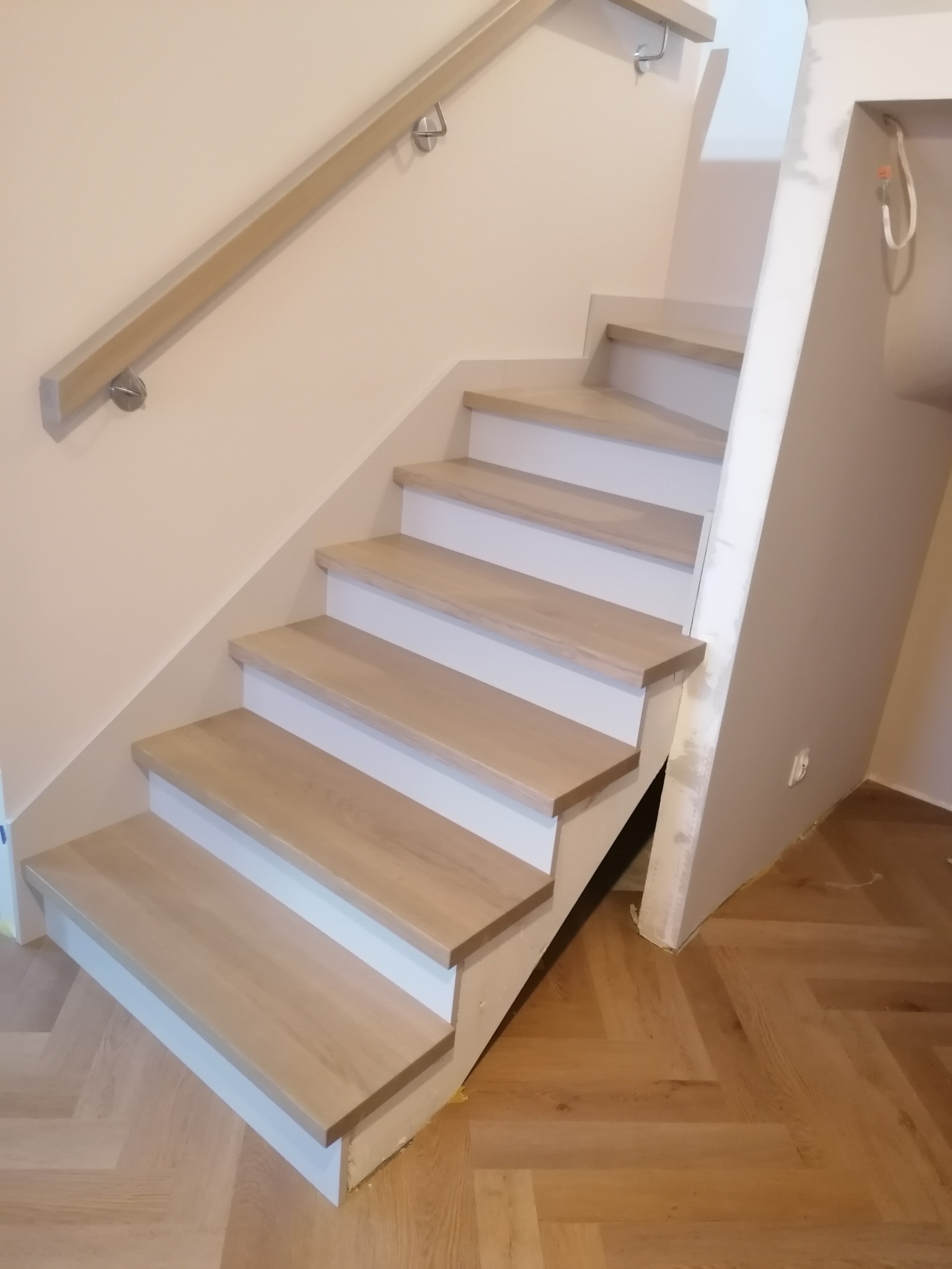 Jasne, drewniane stopnie schodów z białymi podstopnicami i drewnianą poręczą na ścianie, ułożone na parkiecie w jodełkę.