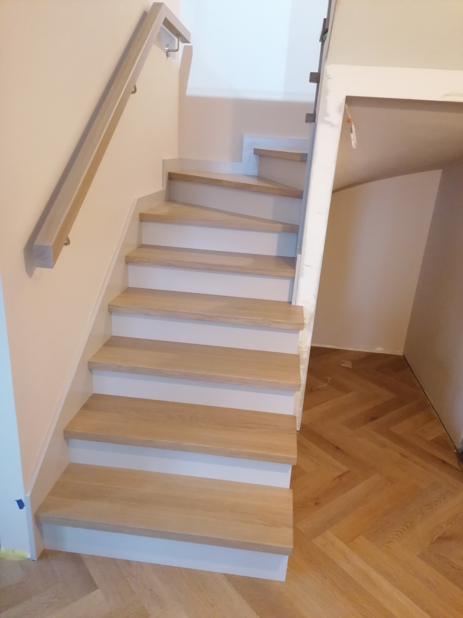 Jasne, drewniane schody z białymi podstopnicami i prostą, drewnianą poręczą, ułożone na parkiecie w jodełkę, widoczne ślady prac wykończeniowych.