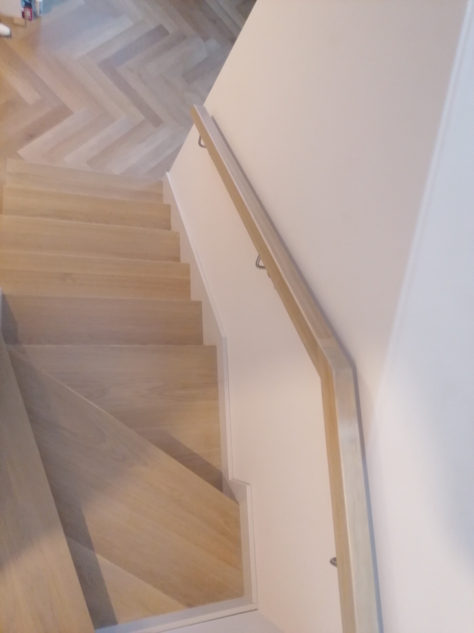 Jasne, drewniane schody z subtelnym wzorem jodełki na podłodze, widziane z góry wzdłuż biegu schodów, z minimalistyczną drewnianą poręczą przymocowaną do białej ściany.