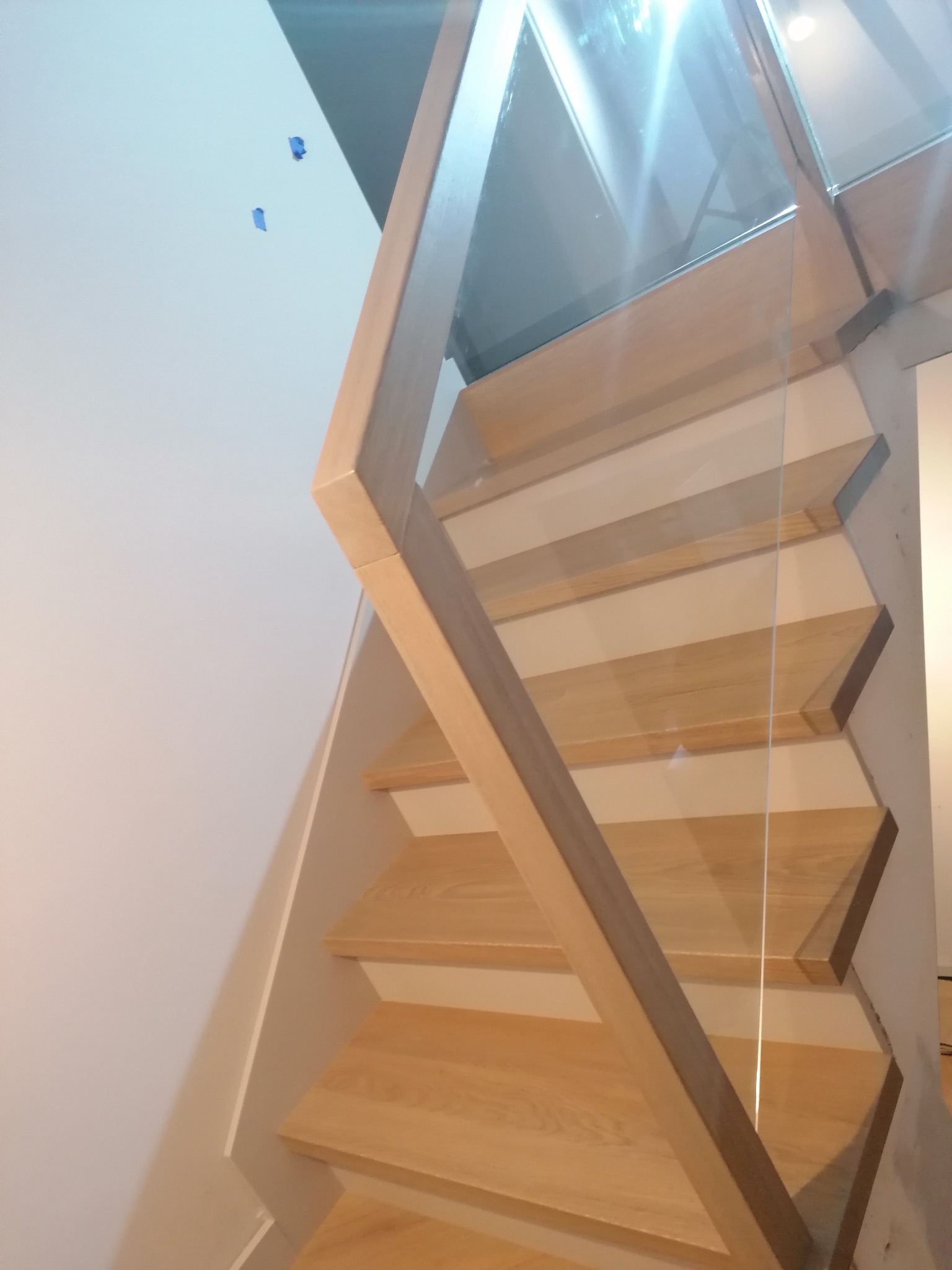 Nowoczesne schody z jasnego drewna z przezroczystą szklaną balustradą, widok z dołu, niebieska taśma malarska na ścianie.