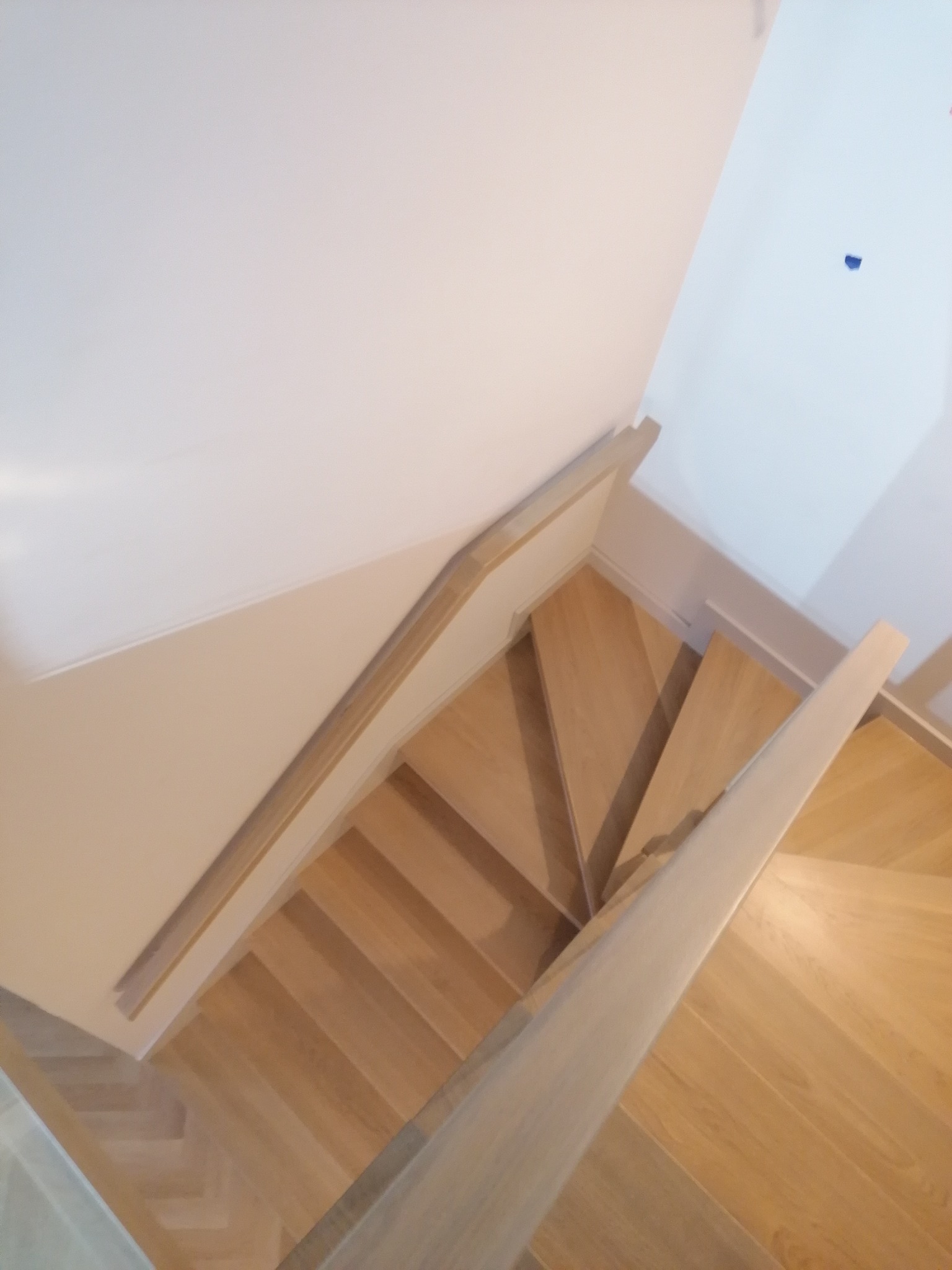 Widok z góry na jasne, drewniane schody z poręczą, ułożone w jodełkę, z widocznym fragmentem białej ściany i sufitu.