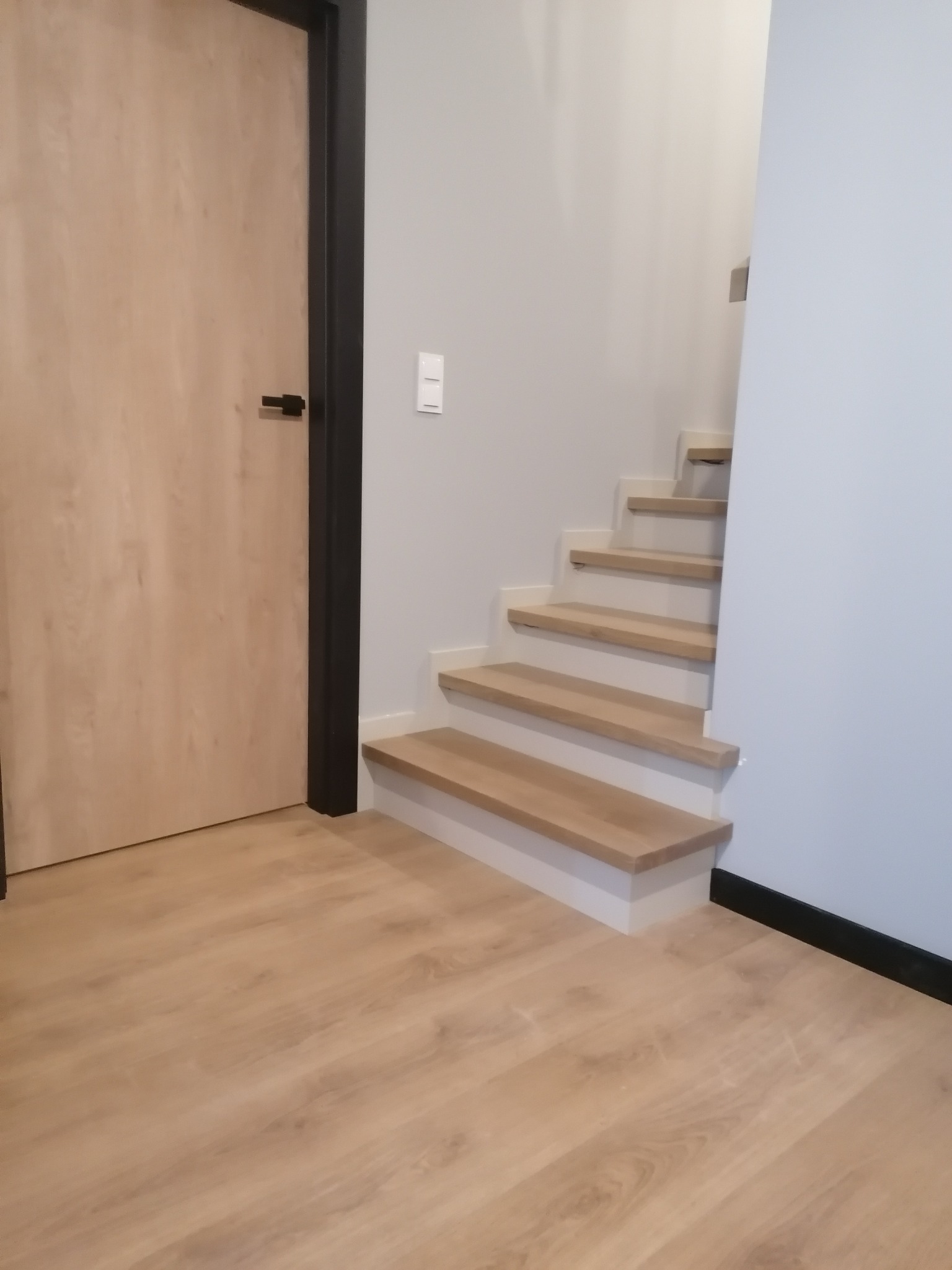 Jasne, minimalistyczne schody z drewna w Dobrzycy, widoczne od dołu, z przylegającą drewnianą podłogą i drzwiami z czarną ościeżnicą.