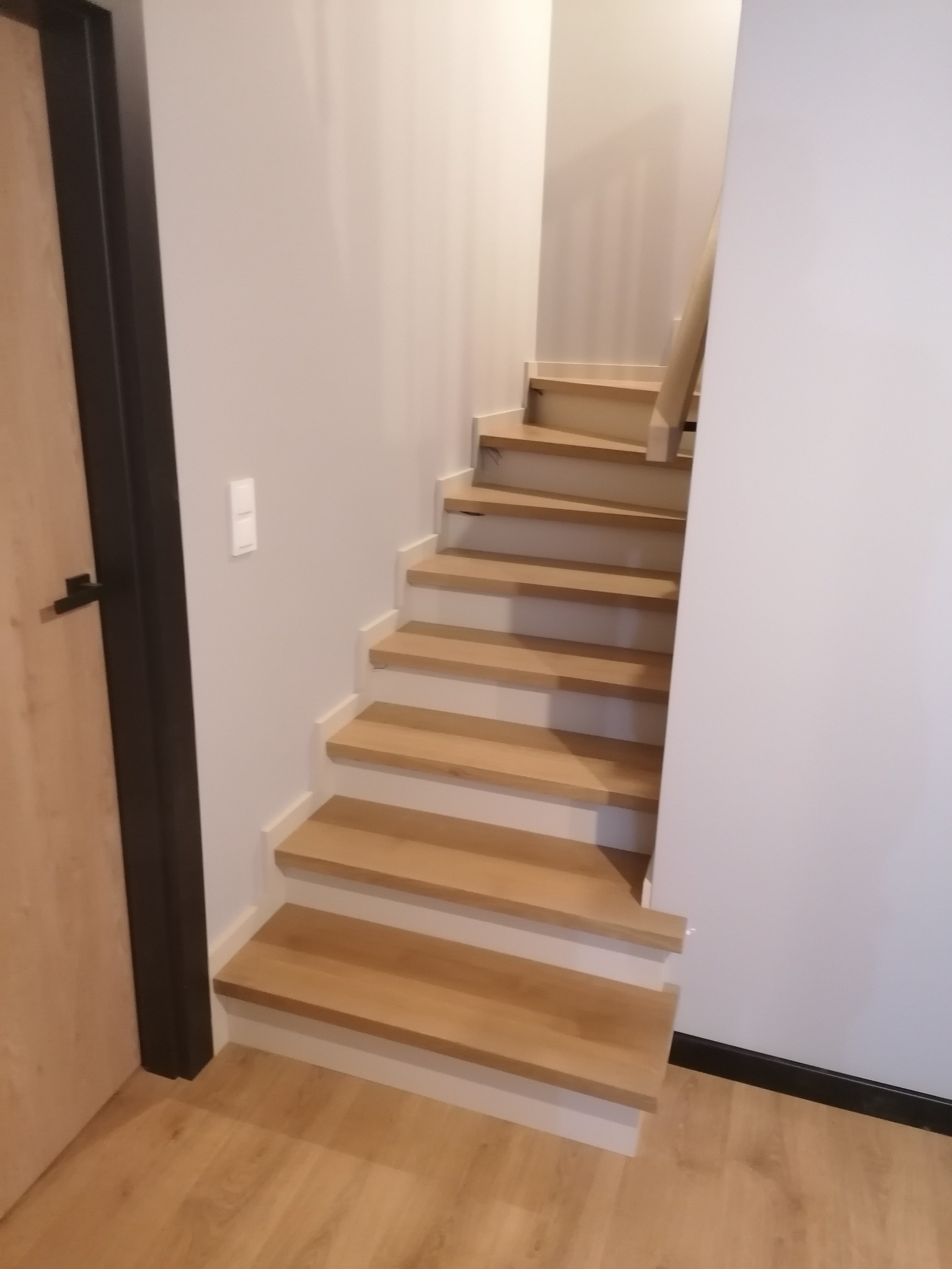 Jasne, drewniane schody wspornikowe z drewnianą barierką, z białymi ścianami i podłogą z paneli w odcieniach drewna, widoczne z poziomu podłogi z perspektywą do góry.