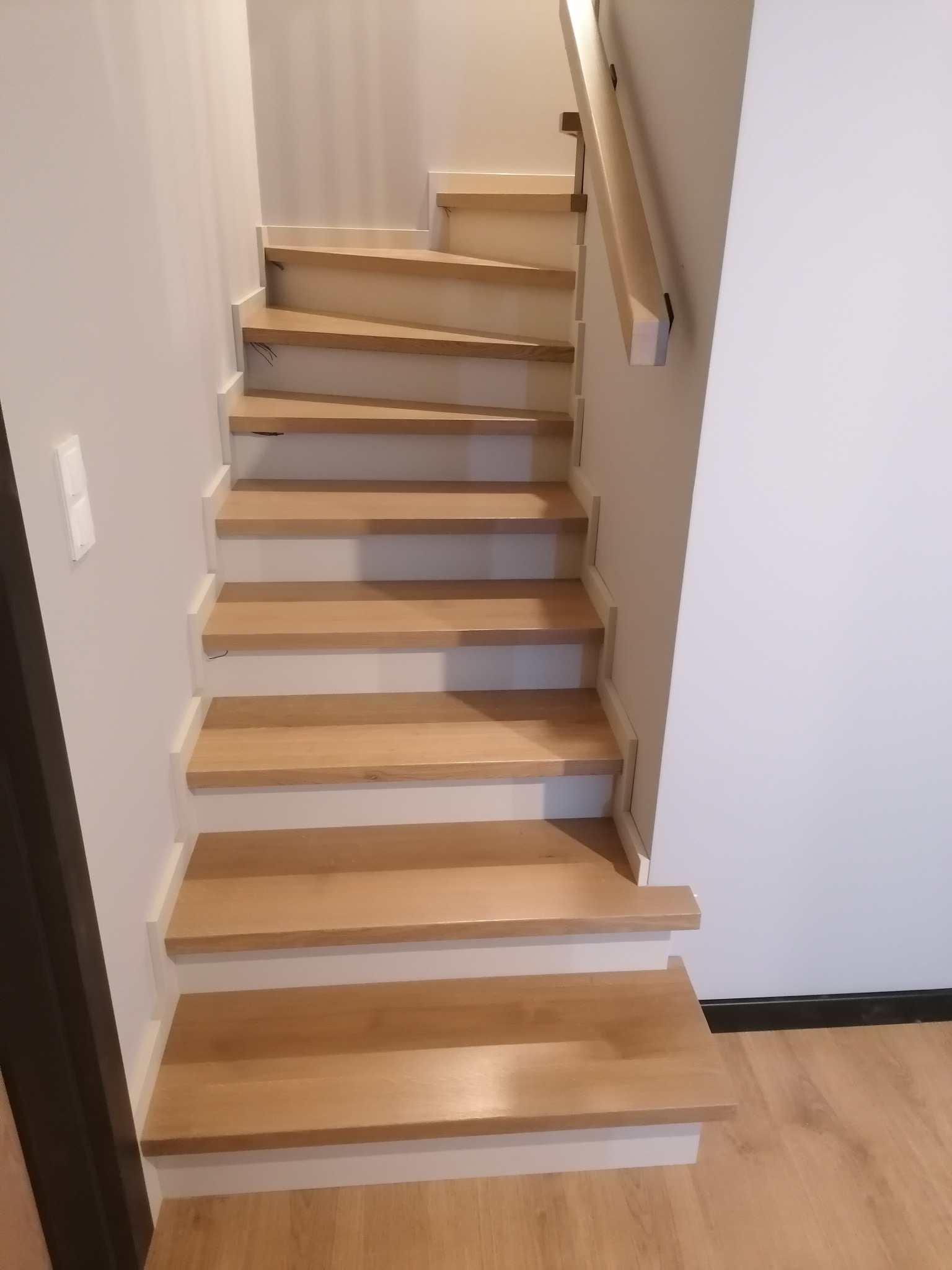 Drewniane schody z jasnego drewna z białymi podstopnicami, proste, bez ozdobników, z drewnianą poręczą po prawej stronie, widok z dołu do góry, na podłodze panele imitujące drewno.