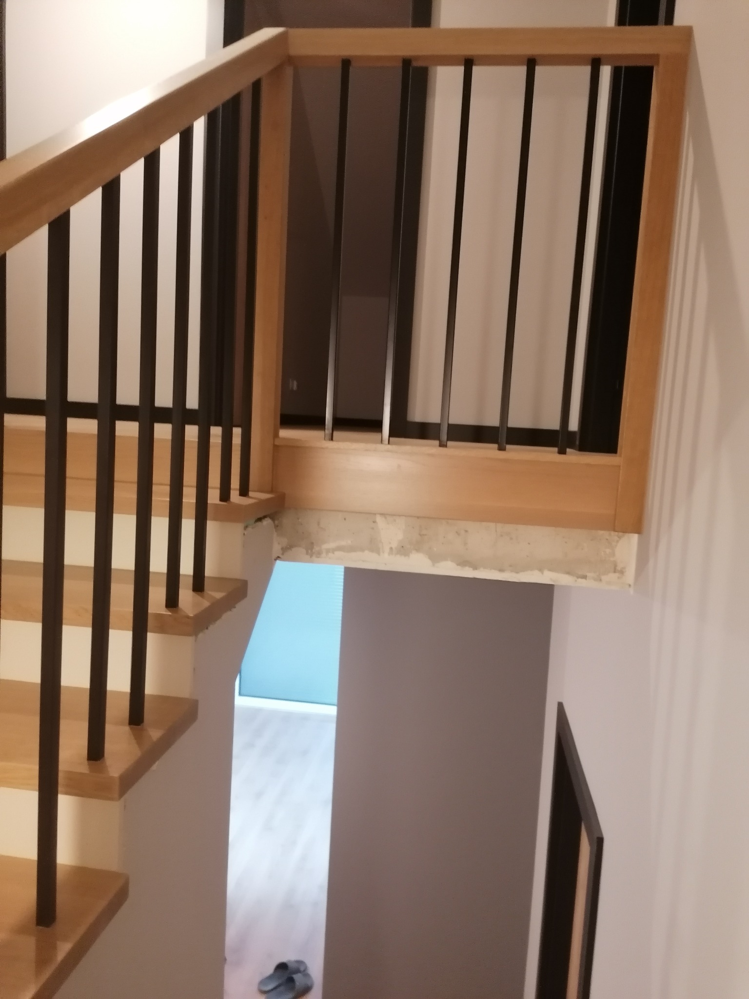 Widok z góry na jasne stopnie z drewnianą balustradą z czarnymi, pionowymi szczeblami, w tle widoczne wejście do pomieszczenia z błękitną ścianą i szare kapcie.