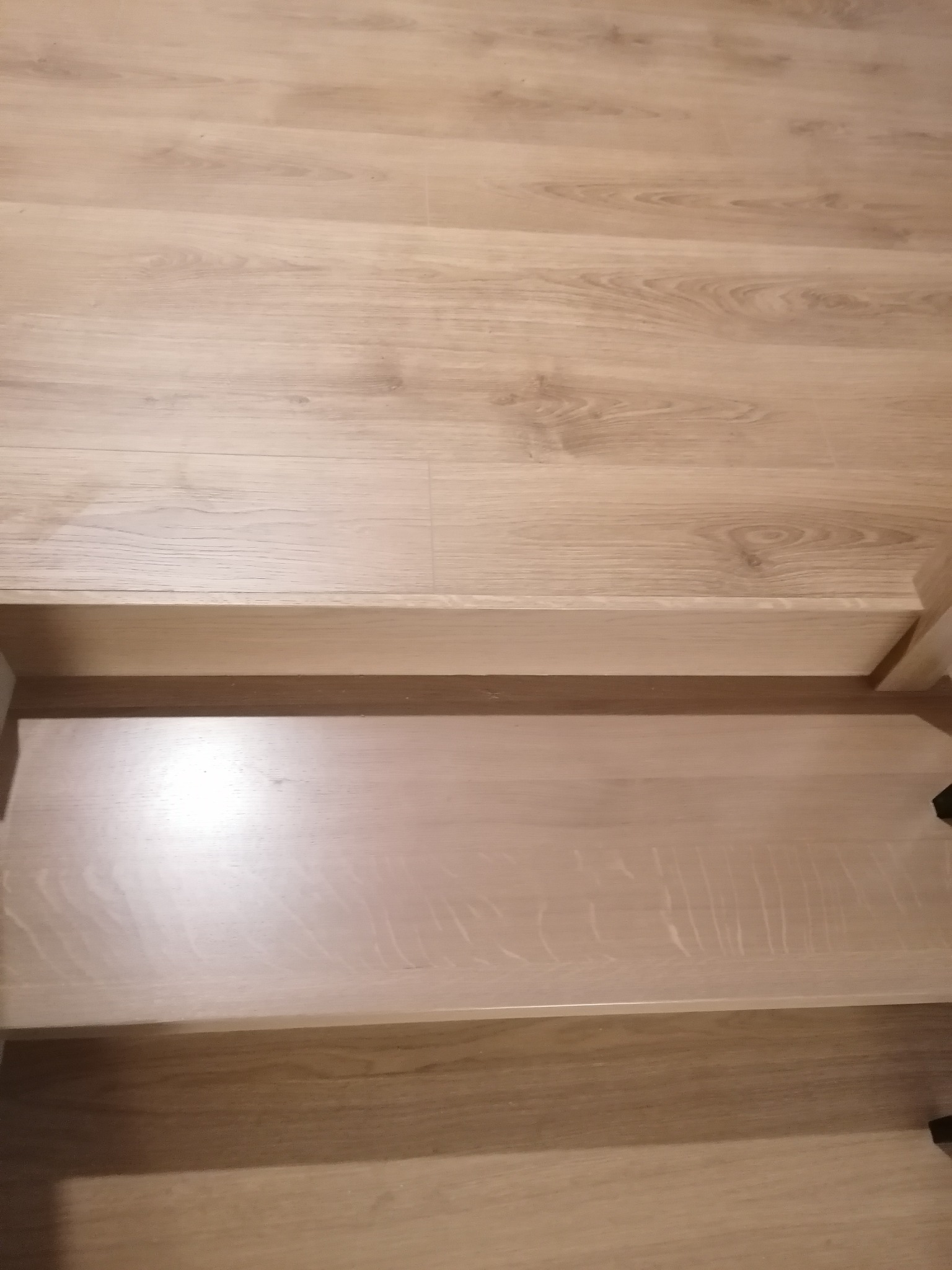 Fragment drewnianych schodów z widocznym usłojeniem drewna i krawędzią stopnia, oświetlonych ciepłym światłem.