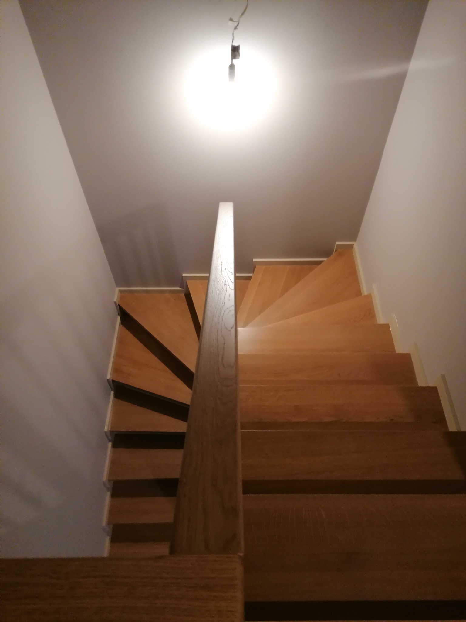 Widok z góry na jasne, drewniane schody spiralne z prostą, drewnianą balustradą, oświetlone pojedynczą lampą sufitową, prowadzące w dół.