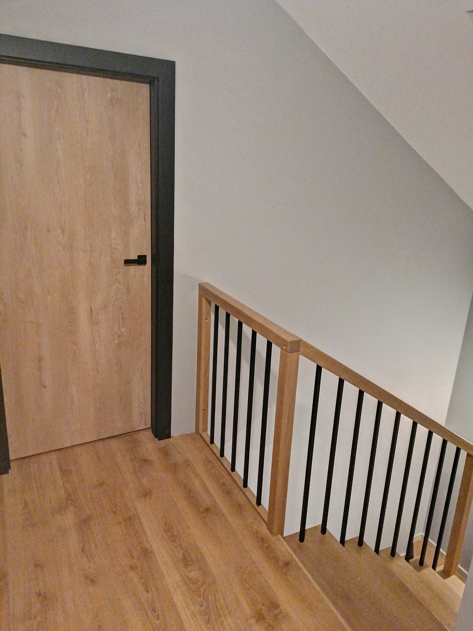 Jasne, drewniane stopnie z balustradą z czarnymi prętami, widok z dołu schodów, obok drzwi z czarną ościeżnicą i klamką.