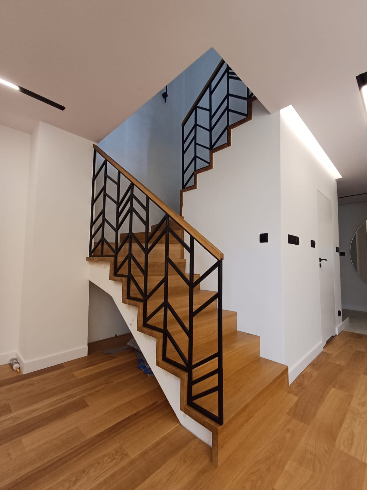 Nowoczesne schody z drewnianymi stopniami i czarną, geometryczną balustradą, oświetlone ledowym światłem sufitowym, widok z dołu na tle białych ścian i drewnianej podłogi.