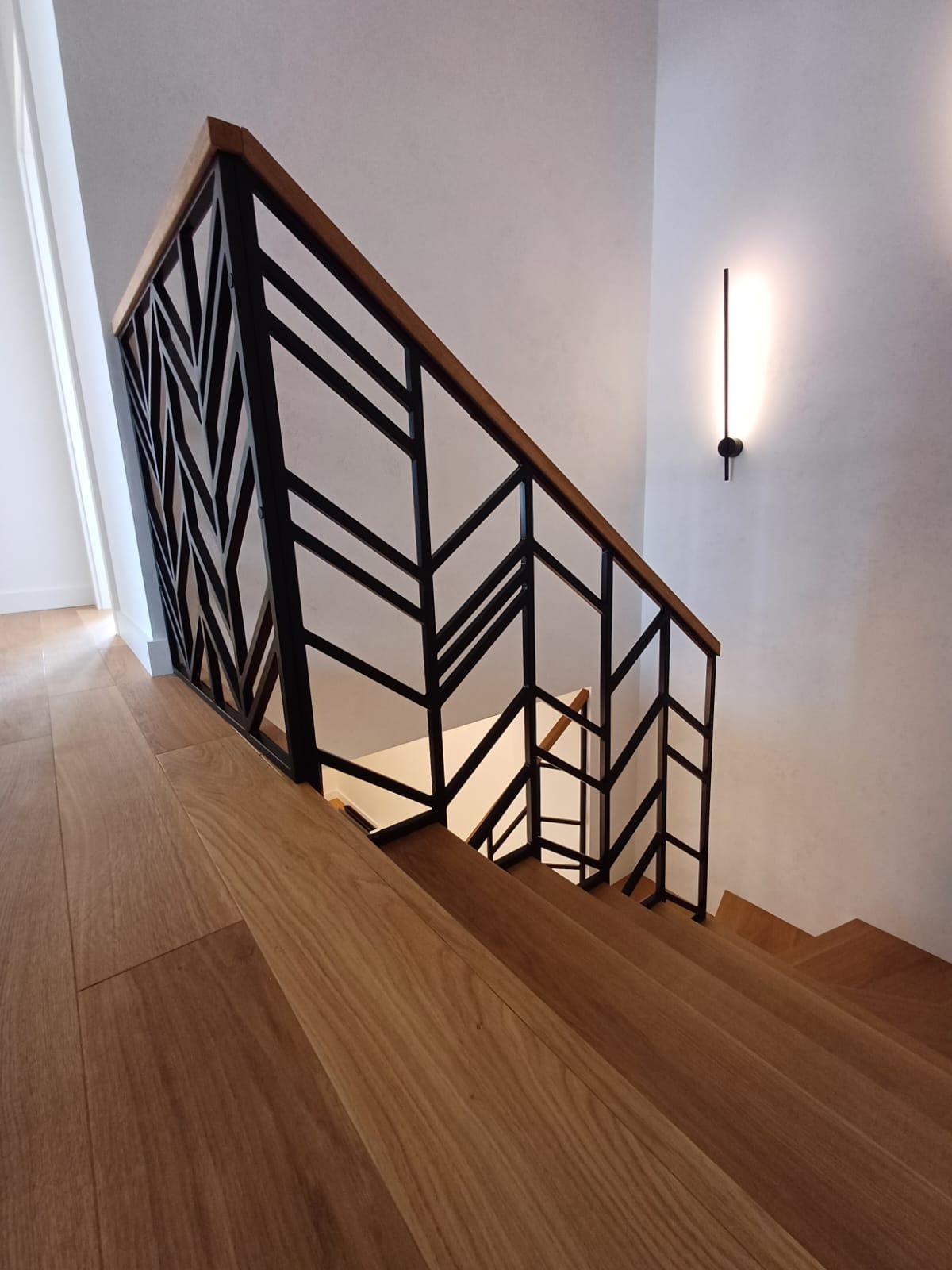 Nowoczesne schody z drewna z czarną, geometryczną balustradą i minimalistyczną lampą ścienną.