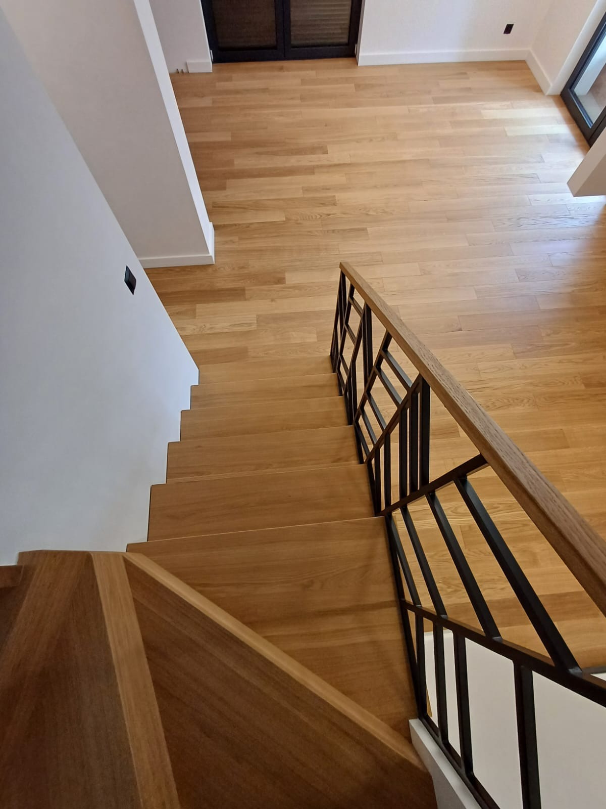 Widok z góry na jasne, drewniane schody z czarną, metalową balustradą o geometrycznym wzorze, prowadzące na drewnianą podłogę w nowoczesnym wnętrzu.