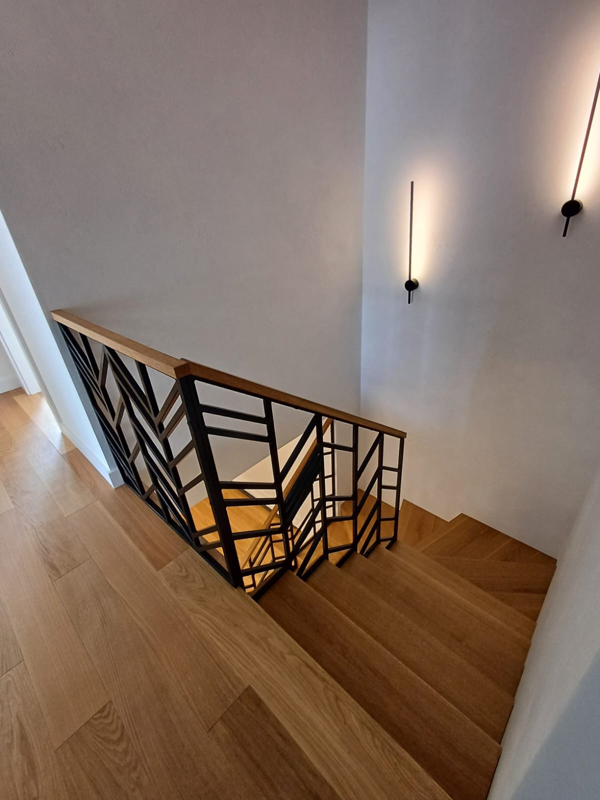 Widok z góry na drewniane schody z nowoczesną, geometryczną balustradą z czarnego metalu i drewnianym poręczą, oświetlone minimalistycznymi lampami ściennymi.
