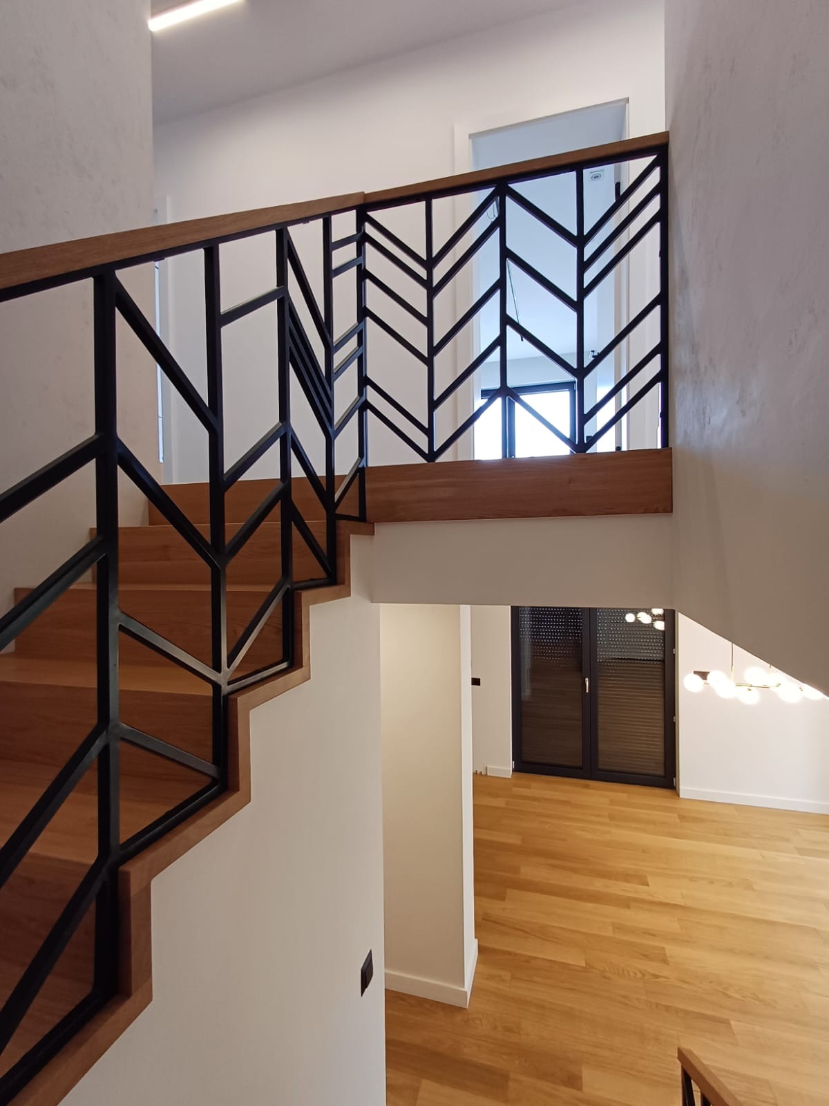Nowoczesne schody z drewnianymi stopniami i czarną, geometryczną balustradą, widok z góry na dół z jasnym parkietem w tle.
