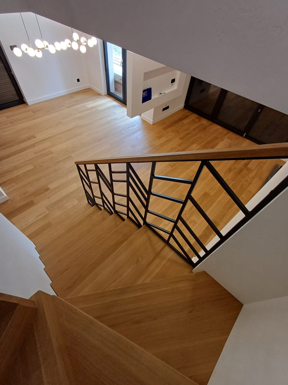 Jasne, drewniane stopnie z czarną, geometryczną balustradą, widok z góry, parkiet w tle, nowoczesna lampa sufitowa.