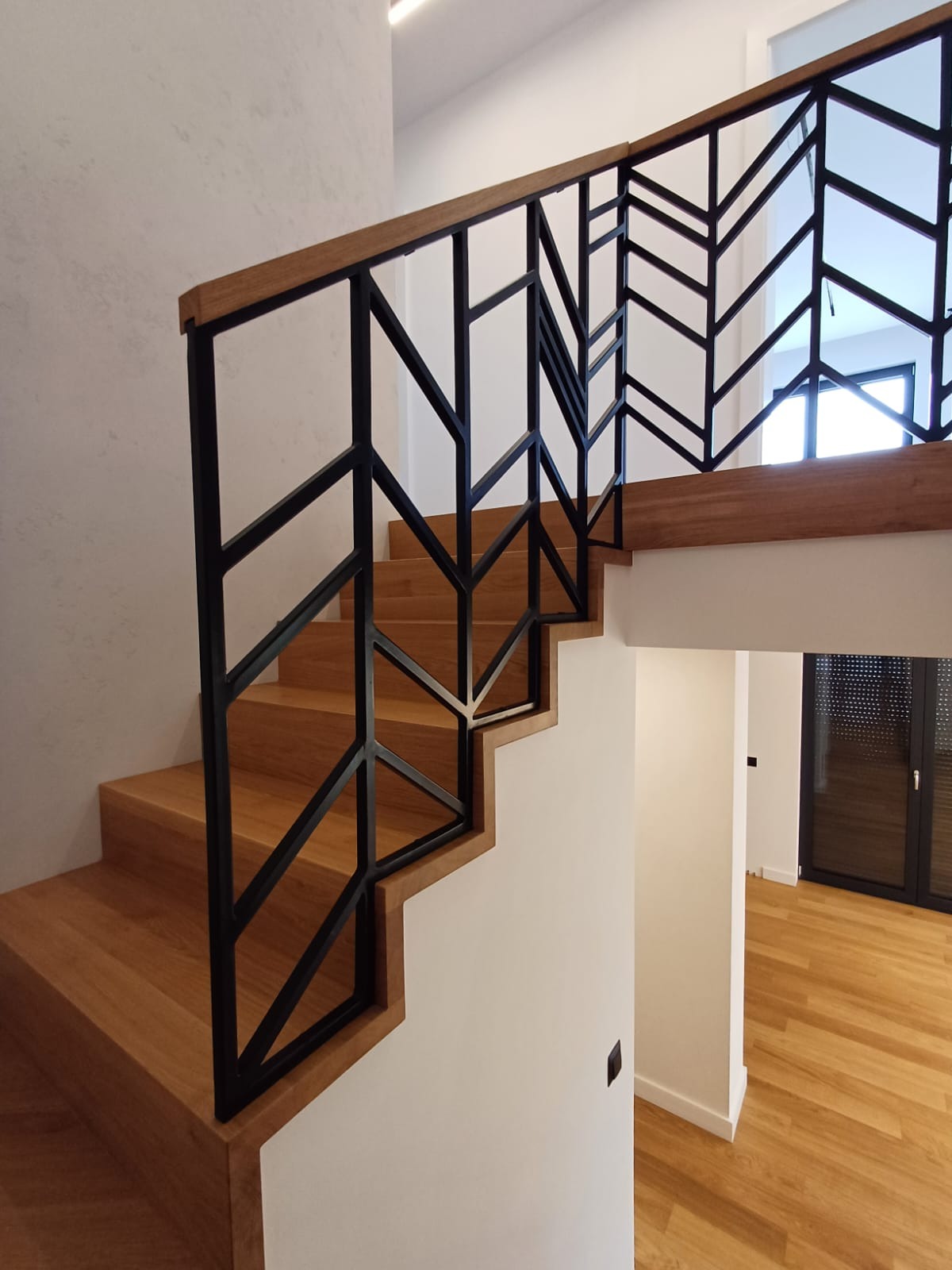 Drewniane schody z czarną, geometryczną balustradą w kształcie strzałek, oświetlone od góry, widok z dołu na podest.