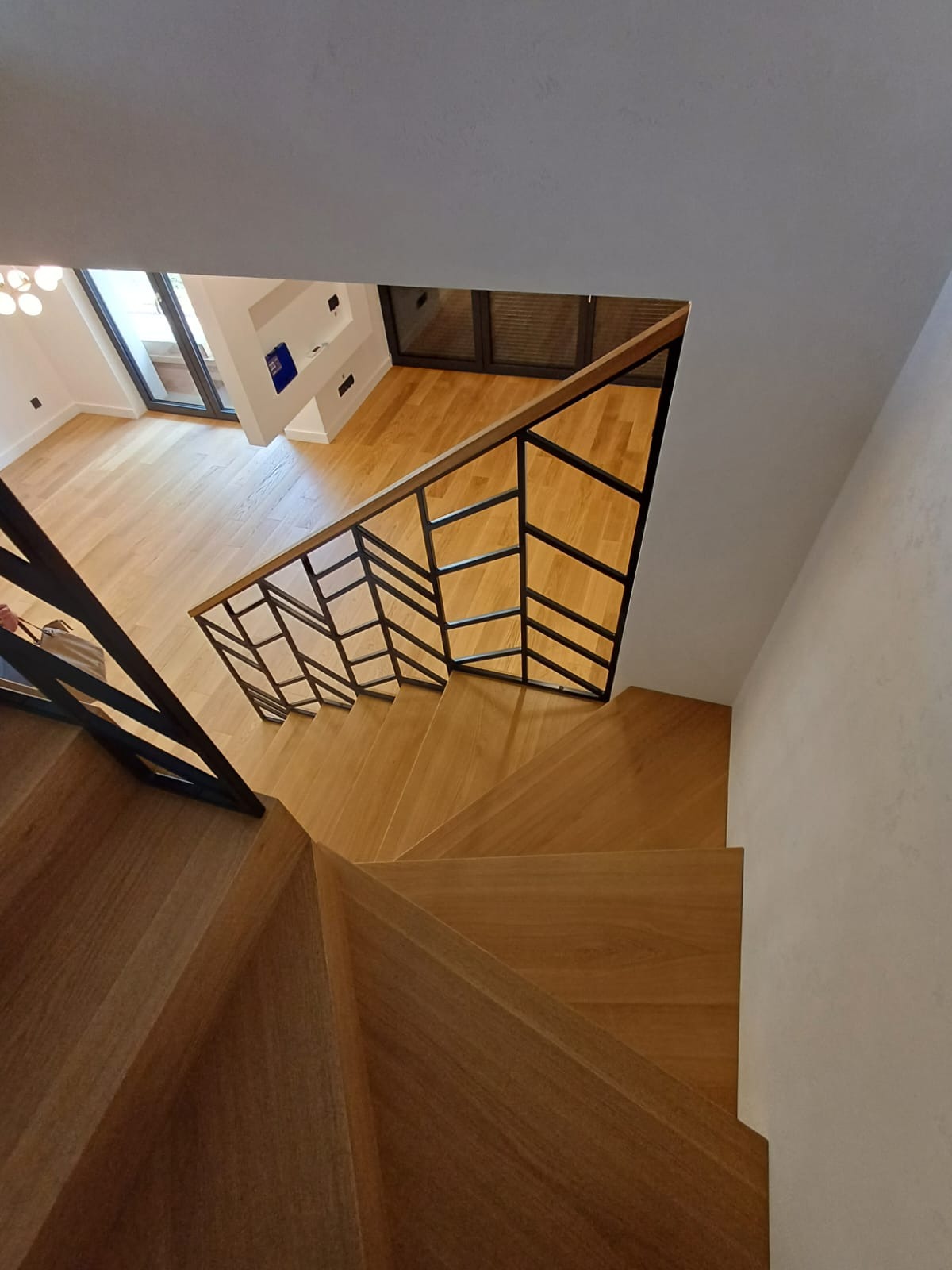 Nowoczesne, dębowe schody z czarną, geometryczną balustradą, widok z góry na parkiet w salonie, jasne wnętrze.