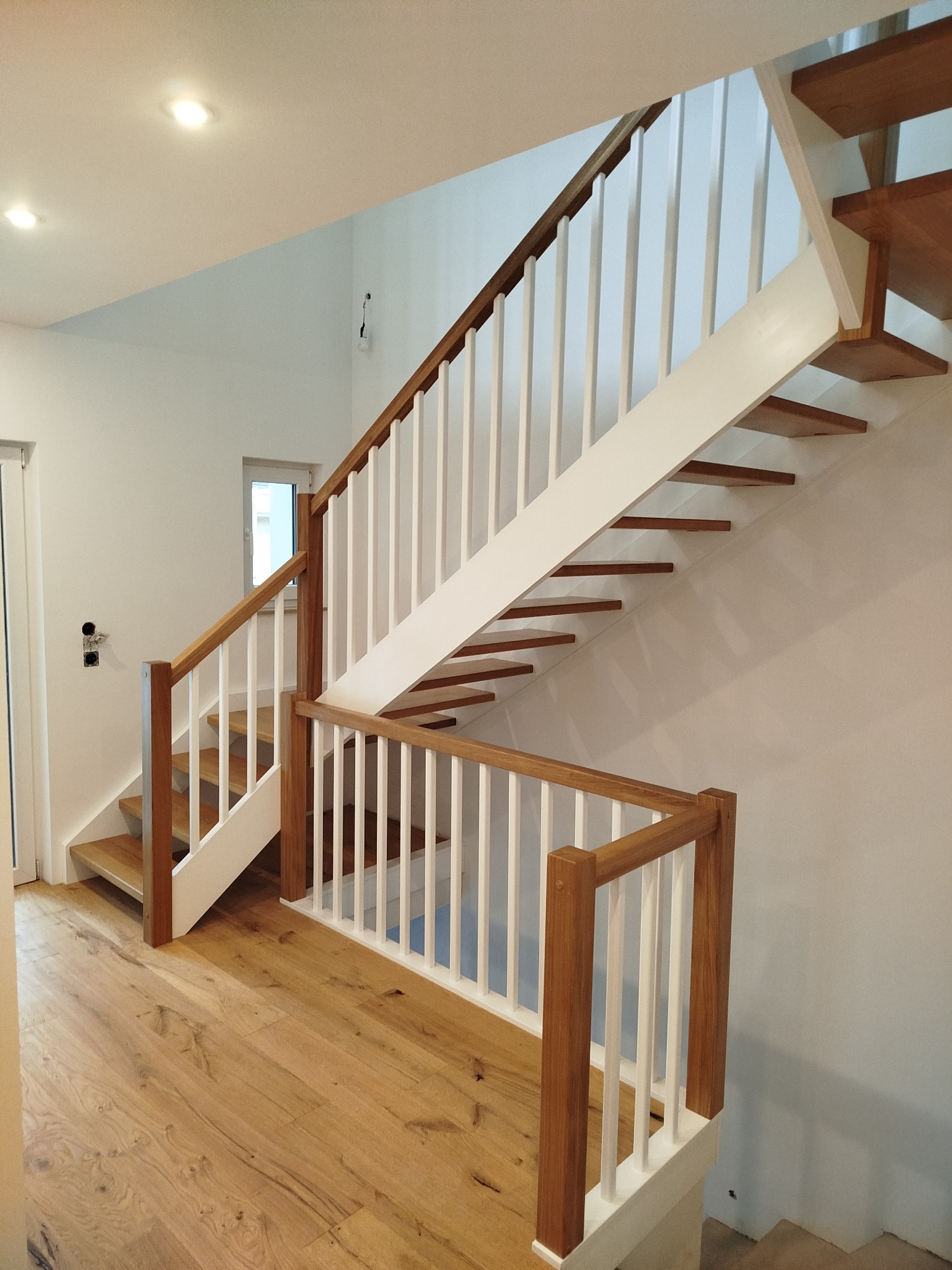 Jasne, nowoczesne schody z drewnianymi stopniami i białymi balustradami, harmonijnie wkomponowane w minimalistyczne wnętrze z drewnianą podłogą.