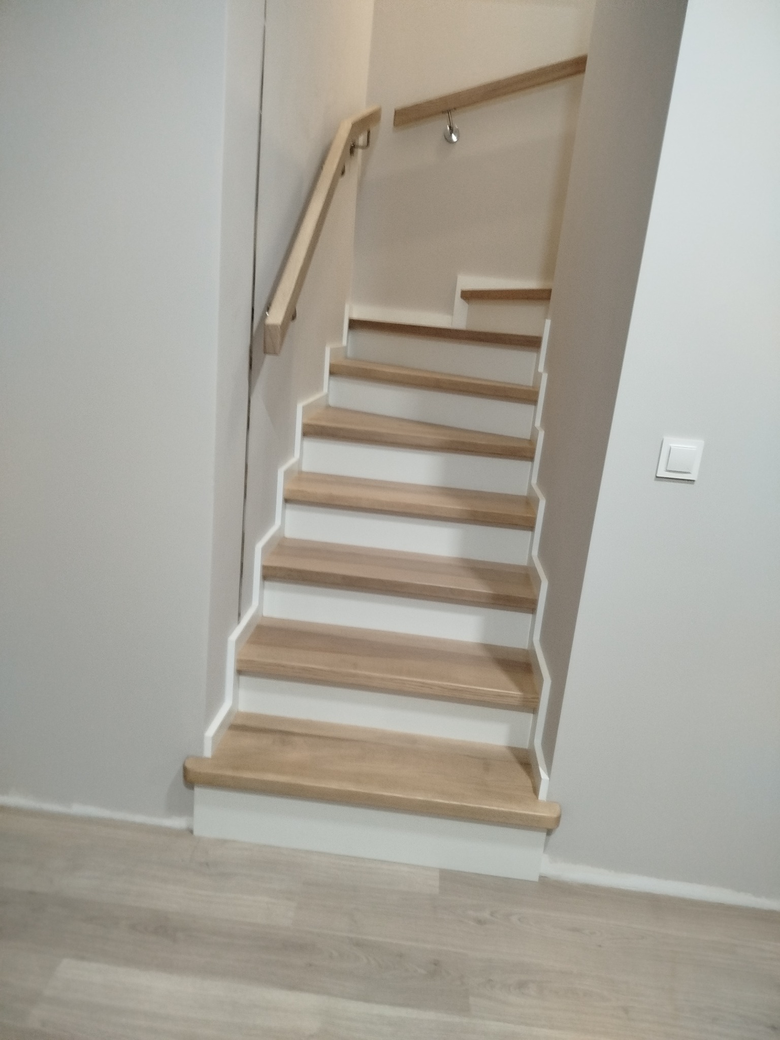 Jasne, drewniane schody z białymi podstopnicami i poręczą, prowadzące w górę. Minimalistyczny design, widok z dołu. Oświetlenie punktowe na ścianie.